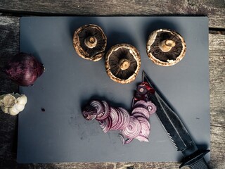 Cutting board on an outdoor wooden camping table with cut red onion, mushroom, garlic, black hunting knife
