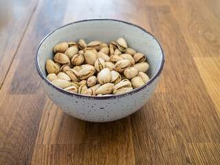 Organic whole pistachio nuts in a white ceramic bowl on wooden table, top view