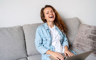 Woman laughing while sits on couch at home and using her laptop.