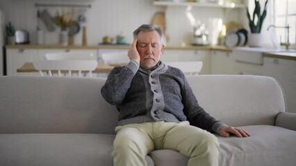 Senior man suffering from terrible headache touching head sitting on couch at home, making massage with closed eyes,doing turning head on sides exercise. Migraine, high intracranial pressure concept.