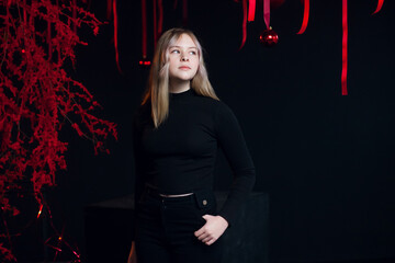 Obraz na płótnie Canvas A beautiful young blonde girl in New Year's black clothes in an atmospheric photo studio with a stylish festive interior in red colors.