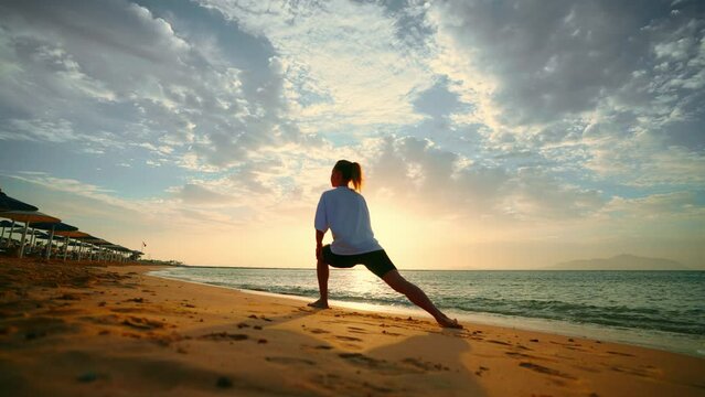 Barefooted woman doing yoga exercise lounge, warrior pose on sandy sea ocean beach at sunset. Female stretching legs. Body care, wellness, healthy lifestyle, sport training physical activity concept.