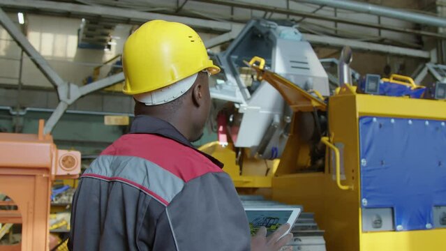 Arc shot of unrecognizable male production engineer in yellow hard hat and coverall holding tablet and comparing caterpillar tractor to its blueprint at machinery plant