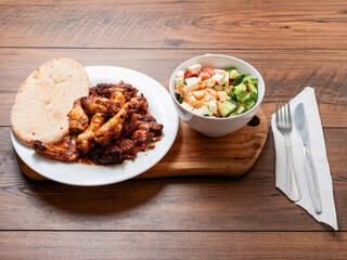 Meal with marinated chicken wings cooked in oven , pitta bread and fresh salad with tomato, cucumber and lettuce on a wooden table.
