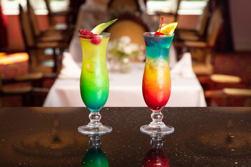 Colorful rainbow layered cocktails garnished with fruit, displayed on a bar counter with a blurred...