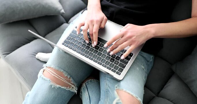 Woman in jeans sitting on sofa at home and typing on laptop keyboard closeup 4k movie slow motion. Freelance and remote work online concept