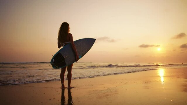 Tourist woman surfer in swimsuit with surfboard stands on ocean sandy beach at sunset admires waves. Summer sport activity on tropics resort. Sporty slim girl. Extreme water sport surfing on vacation.