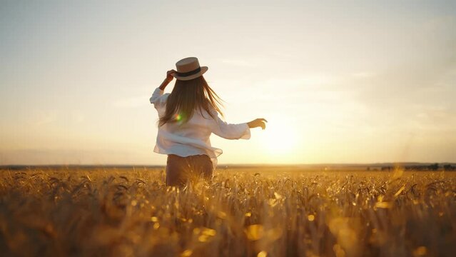 Carefree woman in straw hat running on wheat field feeling freedom, harmony, lightness and serenity, back view. Smiling blonde female enjoying nature resting on farm, outdoors activity. Sunset time.