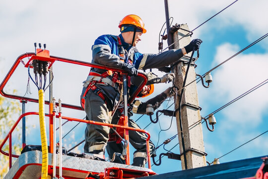 Two professional electricians in hard hats are repairing power lines from cradle of bucket truck. View from below. Electricians change cables on street lighting poles.