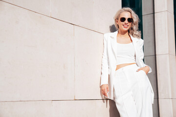 Young beautiful blond woman wearing nice trendy white suit jacket. Smiling model posing in the street at sunny day. Fashionable female outdoors. Cheerful and happy. posing near wall. In sunglasses