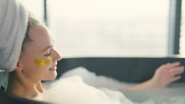 Girl enjoying taking bath with foam. Smiling woman with eye patches, towel on hairs resting relaxing lying in bath looking on pouring water from hands. Home spa wellness self care leisure concept.