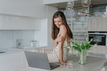 Charming brunette young woman in nightgown sitting on table at kitchen makes video call using laptop toothy smiles enjoying holidays at cozy apartment.
