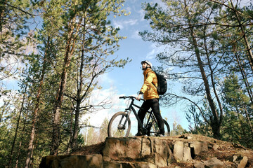 A male cyclist in a protective helmet stands with a mountain bike on a mountain against the sky and enjoys nature