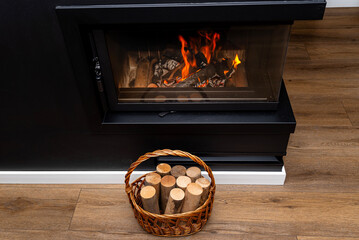 Pieces of birch wood standing in a wicker basket next to a modern fireplace with a closed...