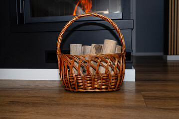 Pieces of birch wood standing in a wicker basket next to a modern fireplace with a closed...