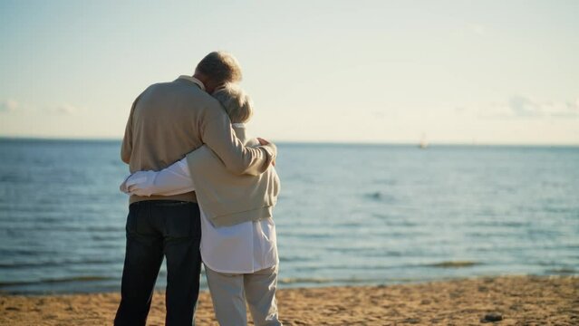Senior couple man woman hugging admiring sea at sunset in evening, back view. Elderly couple family on date cuddling embracing talking. Lifelong relationships happy marriage, love enjoy rest concept.