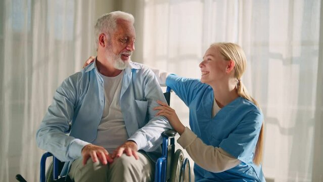 Nurse talking senior man in wheelchair. Female social welfare physical therapist looks after senior male, listening, providing medical care at home. Living with legs paralysis, special needs people.