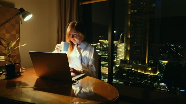 Focused woman working late on laptop in home office with city view. Work-life balance.