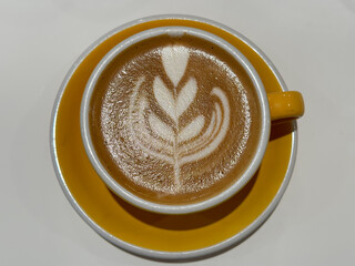 A yellow cup of coffee latte on a white table. A mug of flat white coffee on a white background. Coffee art. Heart flower tulip shape latte art. Copy space. Yellow coffee cup and saucer.