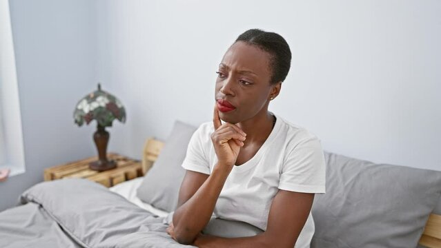 Gobsmacked african american woman lying in bed, awestruck expression painting her beautiful face - hands-on chin in disbelief, feeling a mix of shock and joy indoors in her cozy bedroom.