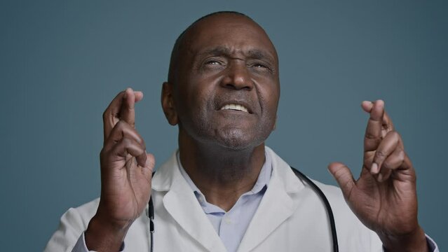 Close-up African American man in studio blue background hopeful male doctor surgeon crosse fingers pray good luck make wish dreaming praying success ask help believe in victory waiting great result