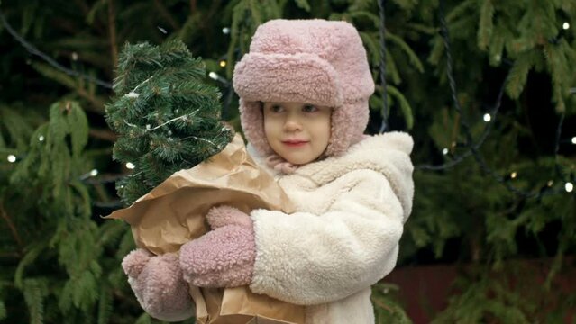A 2-3 year old girl at the Christmas market holding a small Christmas tree. A child catches the falling snowflakes with her tongue.