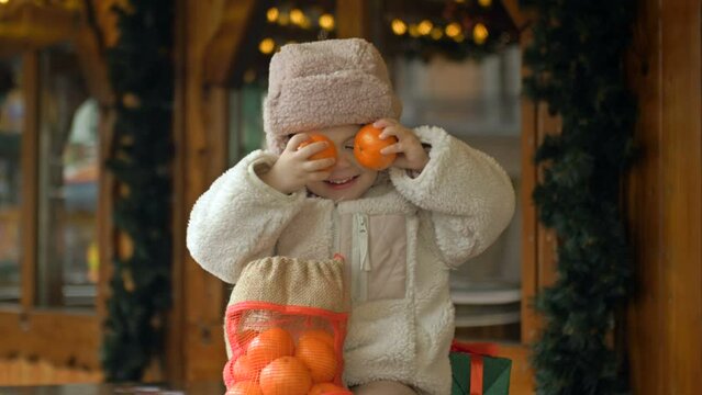 A little girl in warm clothes at the Christmas Fair. The girl jokingly covers her eyes with tangerines.