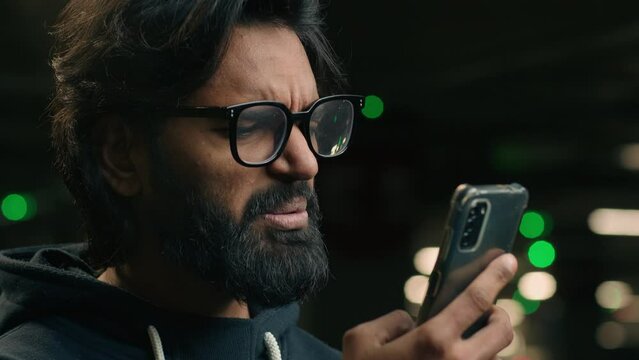 Stressed shocked disappointed sad Arabian man in glasses read bad news in smartphone millennial Indian bearded male hold mobile phone stress by SMS rejection message failure lost crash at parking lot