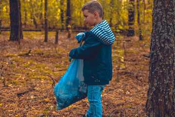 Boy child collects garbage in the park in autumn. A child volunteer with a garbage bag walks around the park and picks up litter lying around. Saving the planet, ecology, pollution.
