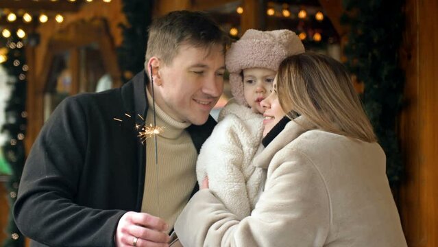 Beautiful young couple with their young daughter at a Christmas fair. The parents hold their baby tenderly in their arms. The adults are holding paper cups of hot drink.