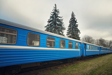 Fototapeta premium Railway, train car riding through city park decorated for Christmas Holidays. Holiday train rides, Christmas tree on background. Christmas train goes through winter forest