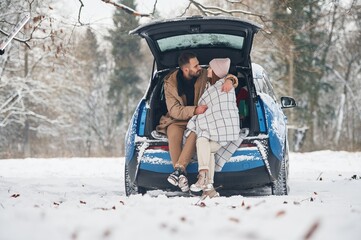 Leaning on the automobile. Happy couple having a walk in winter forest. Blue car is parked