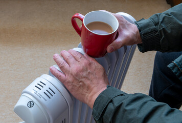 A person holding a hot cup of tea and warming the hands on a portable oil filled heater electric...