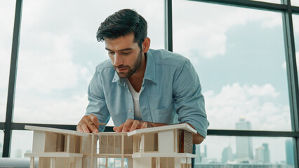 Smart civil engineer measure house model and check, look at blueprint. Professional architect engineer measure, design house model while standing near panorama window with skyscraper. Tracery