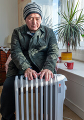 An elderly man fully dressed in the living room sitting next to a portable electric heater trying...