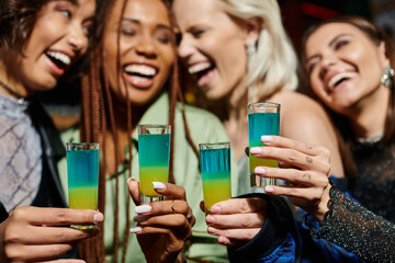 selective focus of shot glasses near cheerful multiethnic girlfriends on blurred background