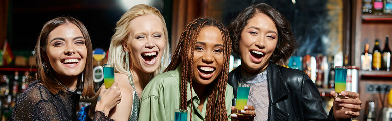 stylish multiracial girlfriends with shot glasses laughing and looking at camera in bar, banner