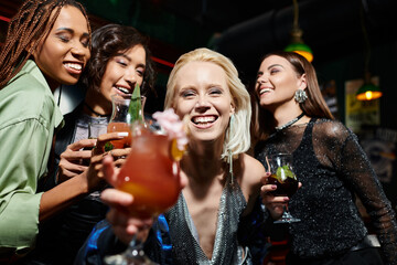 happy blonde woman with glass of cocktail smiling at camera near multiethnic girlfriends in bar