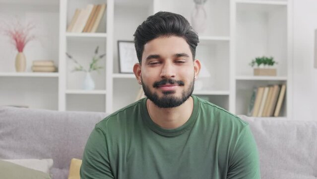Portrait of positive indian man with dark hair and beard resting on comfy grey couch at home. Handsome millennial guy in green t-shirt looking at camera with pleasant facial expression.