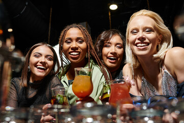 cheerful multiracial girlfriends looking at camera near delicious cocktails in bar, leisure and fun