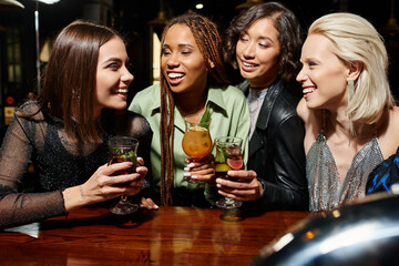 cheerful multiethnic girlfriends with delicious cocktails talking during party in bar, leisure