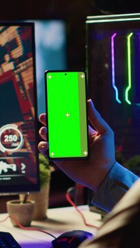 Vertical video Man watching tutorial on green screen phone while playing first person shooter videogame with gun shooting laser bullets. Gamer learning to play game by looking at online guide on
