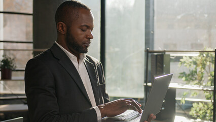 Focused mature adult office worker african american man middle-aged 40s ethnic businessman employee manager CEO specialist stand with laptop typing on computer web surfing internet e-commerce online