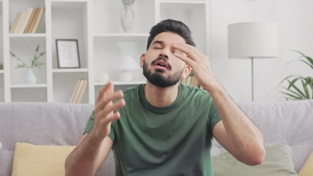 Portrait of disappointed handsome hindu man sitting on grey couch and gesturing with hands raised in shock and disbelief. Negative facial expression and bad news concept.