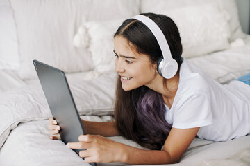 Adorable pre-teen girl in wireless headphones lying on bed with digital tablet device