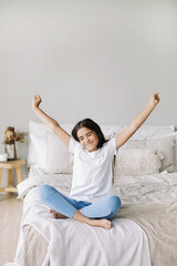 Beautiful pre-teen girl in casual clothes do stretching exercises seated on bed in cozy bedroom in...