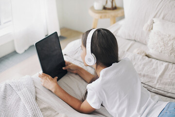 Fototapeta na wymiar Girl in wireless headphones lying on bed with digital tablet