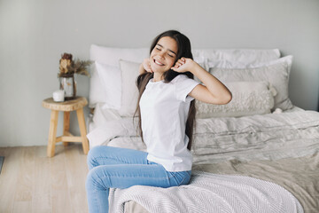 Beautiful pre-teen girl in casual clothes do stretching exercises seated on bed in cozy bedroom in the morning