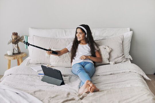 Attractive pre-teen girl resting seated on bed in bedroom holding selfie stick make photos, record video for social media channel, take part in live stream spend free time on internet. Vlogging, fun