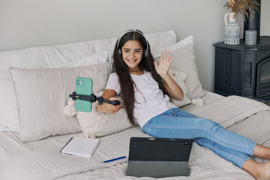 Attractive pre-teen girl resting seated on bed in bedroom holding selfie stick make photos, record video for social media channel, take part in live stream spend free time on internet. Vlogging, fun